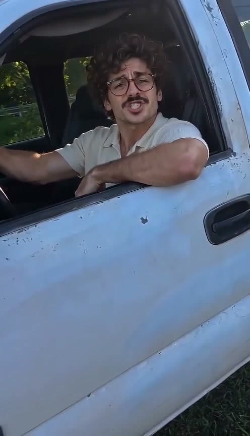 AI-generated man with curly brown hair, round glasses, a moustache and a cream polo gesturing from the open window of a white pickup